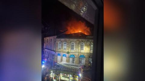 A fire engine parked in front of a building in Kirkby Lonsdale. It is dark and flames are visible behind the building.