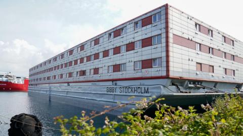 A view of the Bibby Stockholm, a large silver ship with red windows, being used as accommodation barge at Portland Port in Dorset, which has housed up to 500 asylum seekers at a time. In the foreground is a bush and some water.