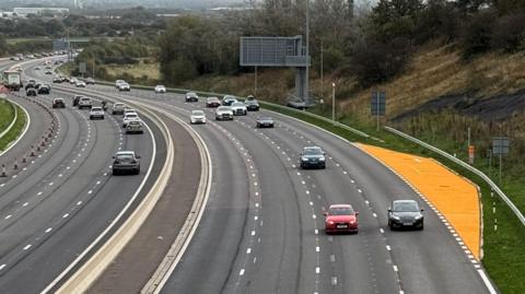 An aerial shot of a stretch of motorway. There are four lanes of traffic heading in each direction. There is also a yellow-painted area in which drivers can pull over in an emergency.