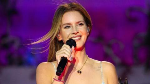 Lana Del Ray smiling as she holds a microphone on stage