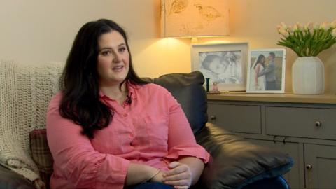 Claire Sugden has long dark hair and is wearing a pink shirt and blue jeans as she talks to the camera. She is sitting on a brown leather sofa with family photographs on the unit behind her.