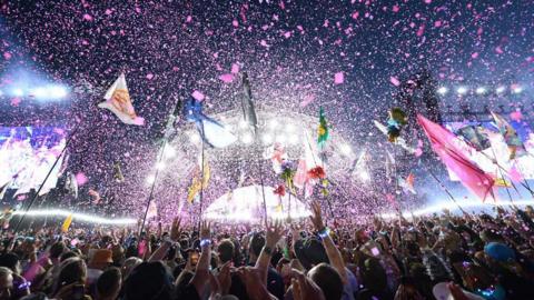 A view of the crowd listening to Coldplay perform on the Pyramid Stage at Glastonbury Festival. There are lots of coloured flags in the air, everyone has their arms up and pink confetti fills the sky and is fluttering down onto the crowd. It is dark but there are very bright lights coming from the stage.