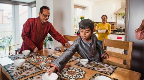 Father and son, in Islamic dress, help set the table while the mother in the background brings a dish