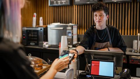 A non-binary person takes payment in a coffee shop via contactless smartphone as they extend the card reader, the customer has blonde hair with pink highlights, and there are coffee cups and coffee machines in the background