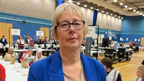 A lady with short, blonde hair, wearing glasses and a blue blazer. She is standing in a sports hall with an election count is happening behind her. 