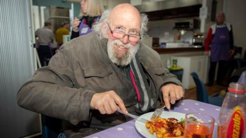 A man smiles at the camera while he sits at a table eating a meal with a knife and fork