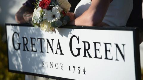 A sign reading Gretna Green since 1754 is leaned on by a woman in a wedding dress holding a bouquet of flowers