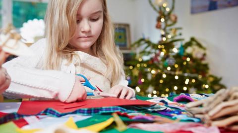 A girl uses scissors to cut colourful paper and materials in a living room, with a Christmas tree in the background.