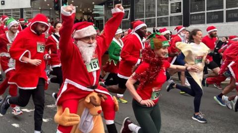 Lots of people in red Santa suits and trainers run along a road. A man with a fake white beard and a Santa hat lifts his hands up in the air.