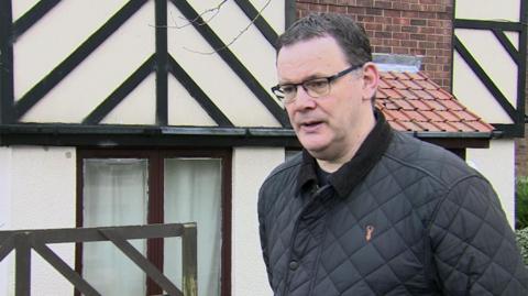 A man with short dark hair and glasses outside a house with Tudor-style features.