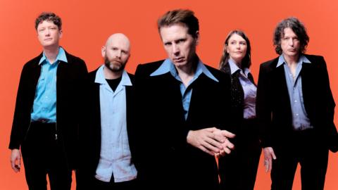 The band Franz Ferdinand (four men and one woman) are looking towards the camera. All are dressed smart casual, with a bright orange background behind them.