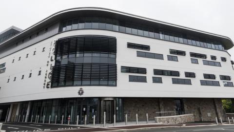 The Jersey Police headquarters in St Helier. A view of the corner of the building showing two sides. The building is curved. The building is cladded in white and has dark windows covered in black metal bars 