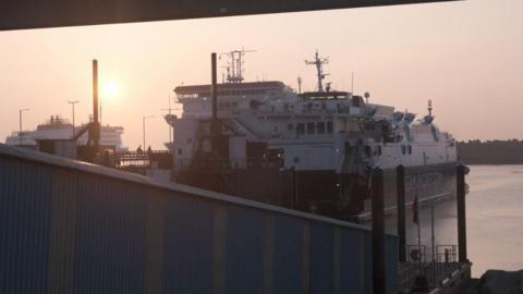 A view of the rear of Isle of Man Steam Packet fast craft Manannan moored in Douglas Harbour with a ramp down to the vessel in the foreground as the sun sets in the background, where another ferry can be seen in the distance. 