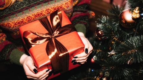 A close up of a person with black painted nailed holding a square gift wrapped in orange paper and a metalic orange bow. They are wearing a striped Christmas jumper made up of warm greens, reds, oranges and yellows standing next to a tree. 