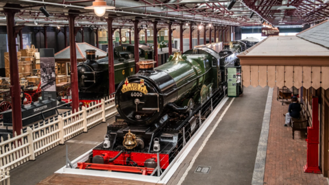 The Bristolian train, which is green and black, inside the STEAM Museum in Swindon. Other items are visible in the background including another steam locomotive