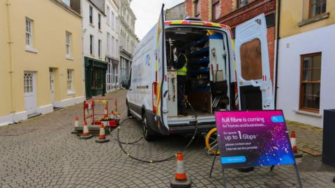 The back of a van which has its doors open and wires coming out into a hole in the ground. There is a sign that reads full fibre is coming in front of it.