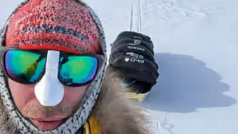 Jonny Huntington wearing a red woolly hat with frost on it, large black-rimmed sunglasses, a white cover on his nose, a head scarf and parka coat while dragging a sled with a large black bag behind him on snow in the Antarctic.