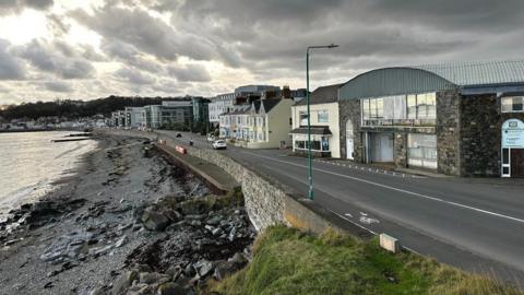 Sea wall with a road going across the top in front of buildings, with traffic passing