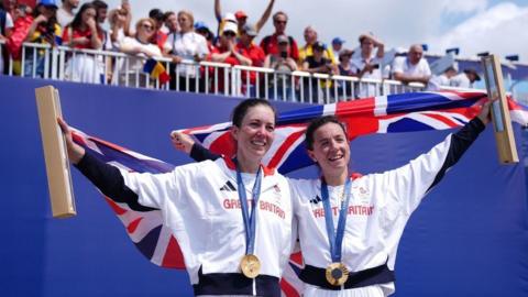 Gold medals hang from the pair's necks as they hold union jack flags behind them with a crowd looking on