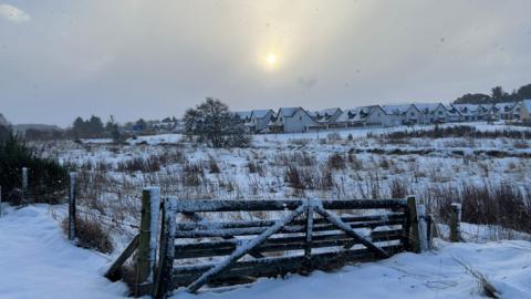 A snowy scene in Aberdeenshire. A farm gate in the foreground is covered with snow. It is mainly brown with snowy patches. In the background, a number of light-coloured houses with snow-covered roofs can be seen.