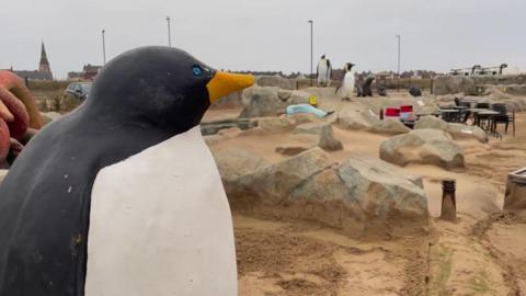 A mini golf course is covered in sand. Various features of the course are peeking out from under the sand including model boulders and large model penguins.