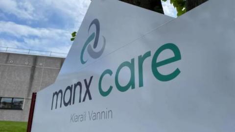 A large white sign with the words Manx Care in English in Manx in grey and green writing on it along with the healthcare provider's logo, which resembles a Celtic knot.