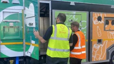 Two men wearing orange and yellow high visibility vests look inside a recycling lorry.