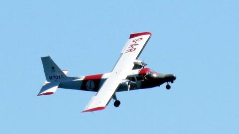 The aircraft, called the Lions’ Pride, airborne. It is white with red writing on the right wing which says 'S-A-R' and has a red block in the middle. The wing at the back has red text on it. The the front there are two windows on the left and is grey.