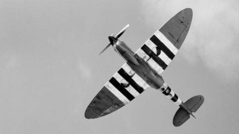 A Spitfire equipped for reconnaissance flight flies overheard in a black and white picture. The aircrafts wings and tail have black and white stripes. 