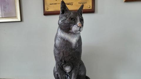 A close-up shot of a life-size statue of a black and white cat against a grey wall, with the bottom edges of plaques and pictures just visible above it