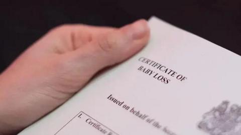 A close-up shot of someone holding a document titled "CERTIFICATE OF BABY LOSS"