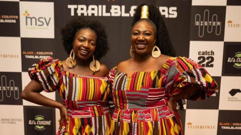 Adeola Adelakun and Ronke Jane Adelakun poses for a photo in front of a  wearing brightly-coloured dresses featuring African-style print. 