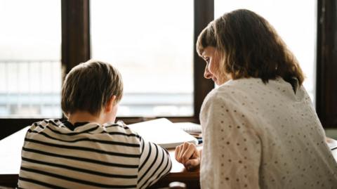 A boy and a woman sit in front of a table - their backs and the side of the woman's face are visible. The boy has brown hair and wears a black and white striped t-shirt, while the woman wears a white blouse. There are notebooks at the table and windows on the back wall.