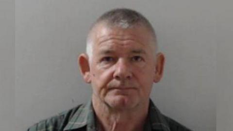 Custody image of Malcolm Sloss, 72, - man with short cropped grey hair, looking into the camera standing in front of a white wall, wearing a green flannel shirt