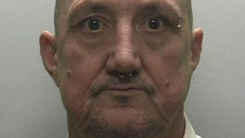 A police mugshot of Simon Mumford. He is a bald man with a septum piercing, an eyebrow piercing on the right side, and light facial hair in the form of a goatee beard. He is wearing a neutral expression.