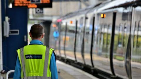 A Southeastern train arriving at a station, as a member of staff in hi vis jacket watches on