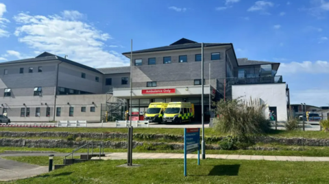 Image shows exterior of emergency department. Two ambulances are parked outside of the large grey building. Green grass banks are in front of the building. 