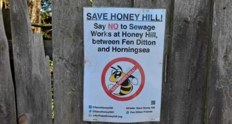 A poster on the wooden fence. The poster reads "Save Honey Hill! Say NO to sewage works at Honey Hill between Fen Ditton and Horningsea". The text is accompanied by an image of a yellow and black bee, with a red circle and line through the image.  