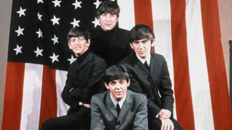 The Beatles, in black and grey suits, shirts and polo neck jumpers, pose for a portrait in front of an American Flag