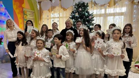 A group of 16 children and two women stand in front of a Christmas tree. The children are dressed in white. A woman stands in the middle of them in a black jumper. They are all smiling and holding cards.