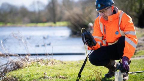 A Thames Water worker, wearing an orange hi viz jacket, is performing test by a river. He's wearing a helmet, gloves and is looking at a testing device. It is a sunny day.