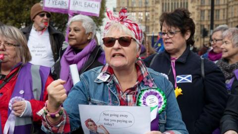 Six women who are part of the Waspi campaign protesting outside Westminster. They are wearing suffragette colours of purple white and green. One woman who is the centre focus of the photo is wearing her short white hair up in a red and white headband with a denim jacket and a checked shirt