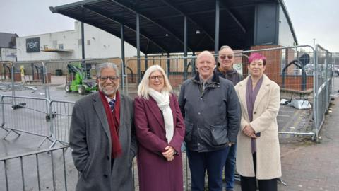 Five people stand in front of railings around a shelter at a bus station. There are three men and two women in the group, all of whom smile at the camera. The silver railings surround a tall black overhead canopy, and a white building with a black Next sign is behind the shelter.