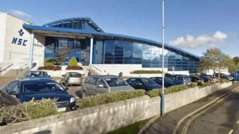 The exterior of the National Sports Centre, which is a large glass-fronted building with a blue sloping roof. NSC is written on a white wall to the left and there are cars parked in front of it.