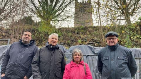 Four people standing in front of metal barriers and granite-filled bags, supporting a collapsed stone wall. St Laurence's Church tower is in the background.