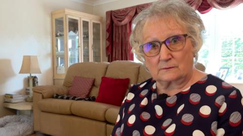 A woman with short grey hair and glasses wearing a spotty blouse, looking straight at the camera with a sofa, a window and a cabinet in the background.