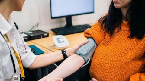 Doctor takes blood pressure of woman sitting with sleeve rolled up. 