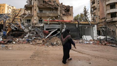 A woman dressed in a black headscarf and dark clothing walks past the wreckage of heavily damaged buildings in Beirut’s southern suburbs, Lebanon. The scene shows extensive destruction, with collapsed walls, twisted metal and debris scattered across the ground. 