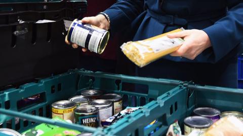 Close up of a woman's hands as she picks out items from crates at a foodbank. She has a tin in one hand and a packet of spaghetti in the other, with more items in front of her. 