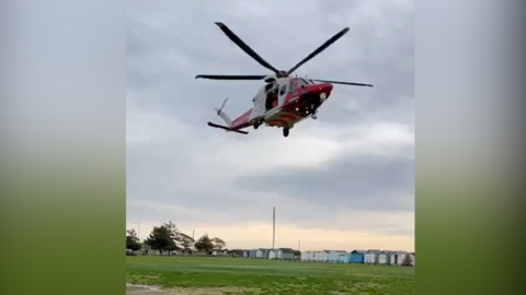 A red and white helicopter taking off from a field with beach huts in the background